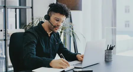 Man behind laptop on phone call on headset taking notes